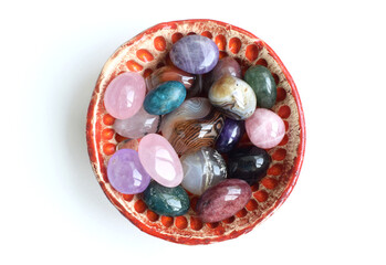 Wall Mural - Colorful tumbled semi-precious stones lie in a handmade ceramic bowl. Red bowl with agate, rose quartz, amethyst and apatite on a white background.