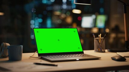Fototapete - Laptop Standing on a Desk with a Green Screen Chromakey Mock Up Display. Table with Computer, Coffee Mug, Headphones and Notebook in Creative Office at Night.