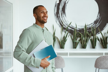 Smiling african businessman holding laptop while standing in cozy office and looking at side