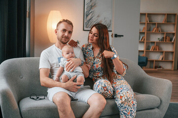 Sitting on the sofa. Happy couple with their newborn boy is at home together