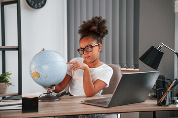 Wall Mural - Examining the globe. Cute female african american student child is sitting by the laptop