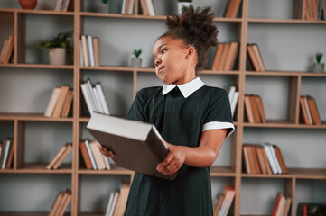 Wall Mural - Holding big book. Cute african american girl in school uniform is at home library