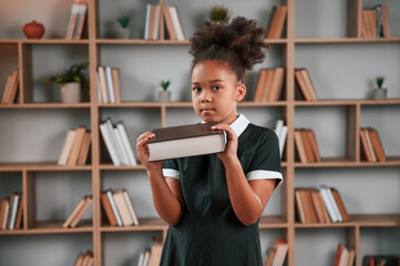 Wall Mural - Holding big book. Cute african american girl in school uniform is at home library