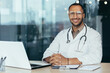 © Liubomir - Portrait of young hispanic male pediatrician doctor. Sitting in the office at a table with documents and a laptop, wearing glasses and a stethoscope in a white coat, looking at the camera, smiling.
