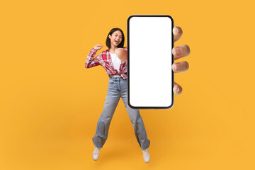 Happy korean lady showing empty smartphone screen while jumping up over yellow studio background, collage, mockup