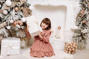 Wall Mural - Adorable child girl with gifts, present boxes near Christmas tree. Smiling kid in a pink dress by decorated fireplace. Happy family at home Christmas New Year December time for celebration concept