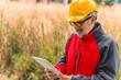 Caucasian, bearded, grey-haired professional construction worker in uniform wearing a bright orange helmet and eyeglasses, standing, holding a tablet used for work, checking information. High quality