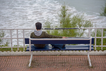 Un hombre joven sentado de espaldas en una banco de la calle mirando relajado el mar en Santander. Persona solitaria descansando en un banco de la calle. 