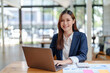 © amnaj - Portrait of smiling Asian businesswoman enjoying her hard work using her laptop at the office and looking at the camera.