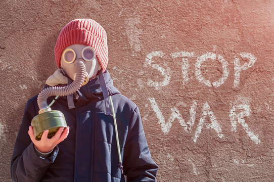 Portrait of a boy in a gas mask and warm clothes against the background of a red old plastered wall. Bright side light. The inscription on the wall stop the war. Concept - no war, world peace