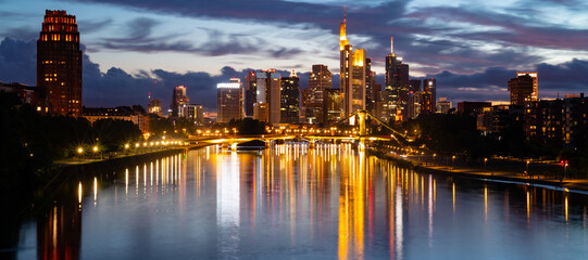 Wall Mural - Frankfurt wide angle nighttime panorama. German city with downtown skyline, tall buildings  and bridge. Evening sky with clouds and reflection in River Main at blue hour with colorful illumination.
