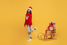 Side View Merry Little Kid Teen Girl 13-14 Years Old Wear Red Xmas Sweater With Deer Santa Hat Posing Carry Gift Boxes On Sled Isolated On Plain Yellow Background. Happy New Year 2023 Holiday Concept.