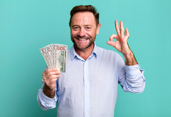 middle age man feeling happy, showing approval with okay gesture. dollar banknotes concept