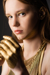 Wall Mural - Portrait of woman with golden paint on hand and face looking at camera isolated on grey