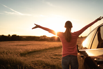 Wall Mural - Feeling happy and free in nature. Young female stopping roadside to watching the beautiful sunrise raising arms outstretched to the sky. 