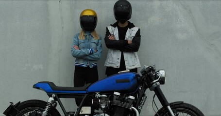 Wall Mural - Portrait of a young stylish couple standing against the wall behind a motorcycle. Loving couple in helmets near the bike