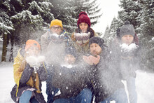 Portrait Of Smiling Young People In Outerwear Walk Together In Snowy Park, Blow Snow At Camera. Happy Friends Have Fun Playing Snowballs On Inter Holiday Or Vacation In Forest.
