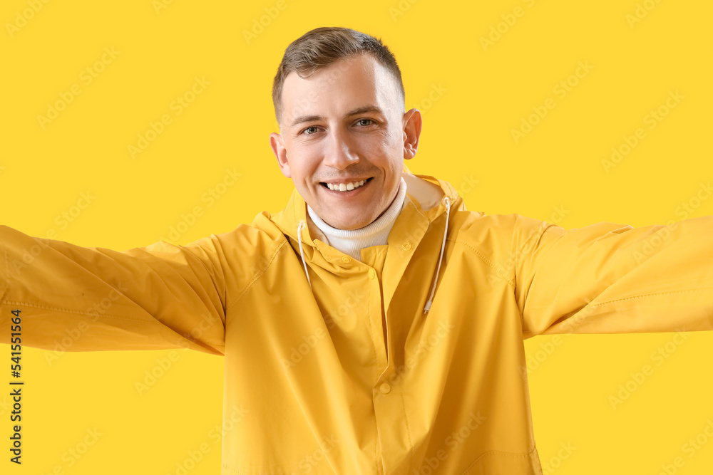 Young man in raincoat taking selfie on yellow background