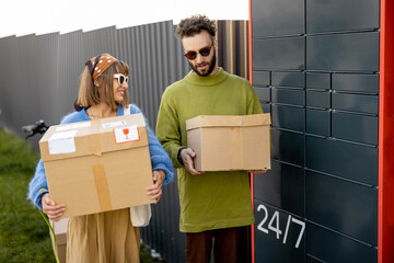 Cheerful young man and woman carry cardboard parcels together received in automatic post machine outdoors. Concept of modern delivery technologies