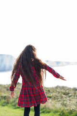 Wall Mural - Little girl with long brown hair in red tartan dress standing on the grass, nature on the background, looking at Cuckmere beach in East Sussex, UK