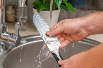 A man washes a sharp steel dirty kitchen knife with a sponge with detergent in the kitchen sink under running water. Gentle hand washing of the kitchen knife. Cleaning of dirty sharp metal products.