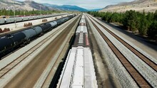 Forward Flight Dolly Drone Shot Flying Over A Cargo Train On A Railroad Station In A Desert Environment On A Sunny Day Next To A Mountains In The Background And Powerines In The Picture An Tank Trains