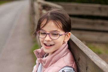 Little beautiful cute girl in glasses smiling leans on a wooden fence. Village child in a vest on a walk in the countryside