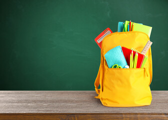 Canvas Print - Backpack with school stationery on wooden table near green chalkboard, space for text