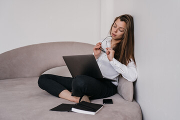 Exhausted  beautiful brunette business woman sitting on sofa with laptop touching head eyes closed leaning on wall. Tired hispanic woman overloaded after hard work. Remote working girl with headache.