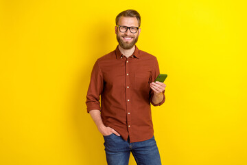 Portrait of cheerful funny glad man with masculine beard burgundy shirt hold smartphone arm in pocket isolated on yellow color background