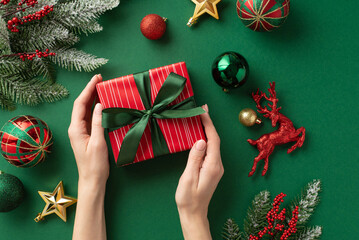 Wall Mural - Christmas concept. First person top view photo of female hands holding red giftbox with bow over green gold red baubles star deer ornaments mistletoe fir branches in snow on isolated green background