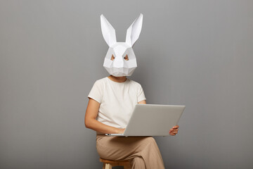 Portrait of anonymous female wearing rabbit mask and white t-shirt, sitting on chair with notebook in hands, freelancer working online, isolated over gray background.