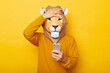 Photo of sad upset male wearing lion mask and orange sweatshirt posing isolated over yellow background, using mobile phone, having problems with internet connection, showing facepalm gesture.