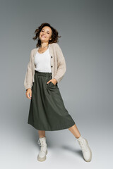 Sticker - cheerful woman in warm cardigan and boots posing with hand in pocket of green skirt on grey background