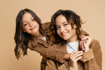 brunette girl in trendy autumn jacket holding hands of mom smiling at camera isolated on beige