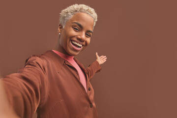 Wall Mural - Young cheerful casual African American woman invites you to follow together smiles and stretched out hand filming himself on phone or camera posing on isolated brown background. Follow me concept