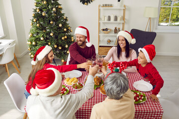 Happy joyful dad, mum, grandma, grandpa and little children in funny hats having Christmas family dinner, sitting around table at home, enjoying meal, eating roast chicken and drinking juice together