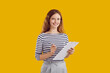 © Studio Romantic - Studio portrait of female secretary or business assistant. Happy beautiful young woman standing isolated on yellow background, holding pen and clipboard, looking at camera and smiling