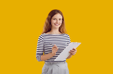 Studio portrait of female secretary or business assistant. Happy beautiful young woman standing isolated on yellow background, holding pen and clipboard, looking at camera and smiling