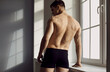 Back view of a male model posing in underwear. Backview of a young man with a beautiful, athletic body, wearing comfortable black underpants standing near the window in the studio. Fashion concept