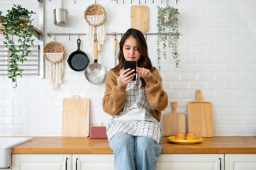 Wall Mural - Happy young asian woman relaxing at home she is sitting on counter kitchen and using mobile smartphone