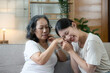 © wichayada - Happy adult granddaughter and senior grandmother having fun enjoying talk sit on sofa in modern living room, smiling old mother hugging young grown daughter bonding chatting relaxing at home together