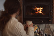 Young woman sitting at home by the fireplace with a mug of hot coffee or tea and a book, she is wearing woollen sweater. Cold houses with low temperatures in Europe concept. Selective focus on a mug.