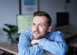 © boytsov - Attractive bearded male sitting in home office looking at camera smiling. Young handsome man in denim shirt, positive emotion with computer monitor on background. High quality image