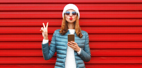 Wall Mural - Portrait of beautiful young woman with smartphone blowing her lips sends air kiss wearing white hat on red background