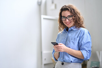 Smiling young business woman user, happy businesswoman wearing glasses holding cellular smartphone working standing in office using mobile cell phone working on cellphone looking at camera.