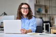 © insta_photos - Young happy business woman company employee, marketing manager sitting at desk working on laptop. Pretty female professional worker using computer in corporate modern office looking away and smiling.