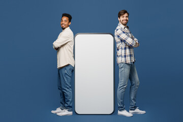 Wall Mural - Full body side view young two friend men wear white casual shirt together stand near big huge blank screen mobile cell phone smartphone with mockup area isolated plain dark royal navy blue background