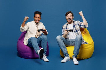 Full body young two friends happy men wear white casual shirts together looking camera hold in hand play pc game with joystick console do winner gesture isolated plain dark royal navy blue background.