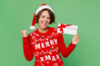 © ViDi Studio - Merry young woman wear xmas sweater Santa hat posing hold store gift certificate coupon voucher card do winner gesture isolated on plain pastel green background. Happy New Year 2023 holiday concept.